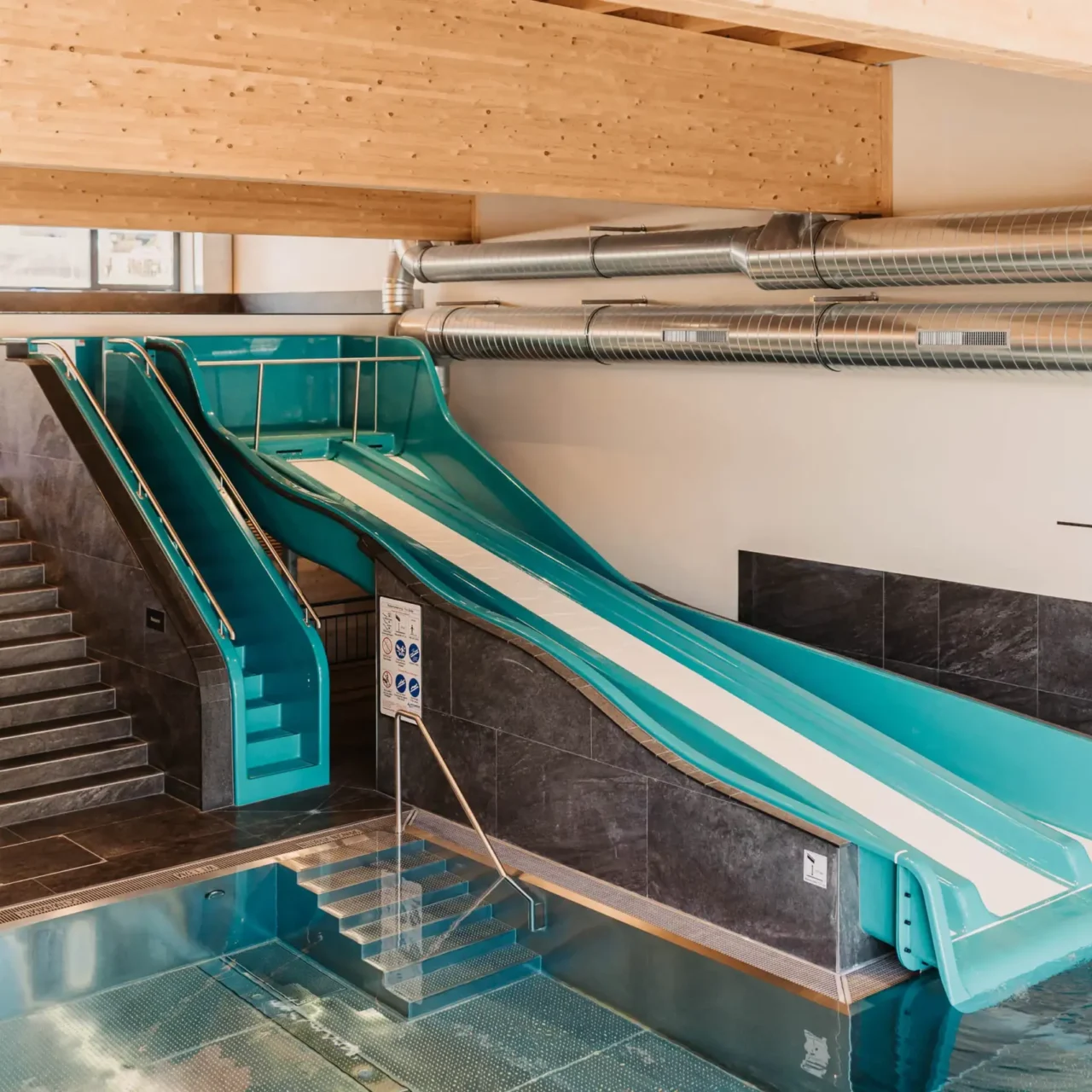 Innenbereich eines Familienpools mit großer blauer Wasserrutsche und moderner Ausstattung – breiter Rutschkanal endet im beheizten Becken, umgeben von stilvollem Design und Holzdecke.