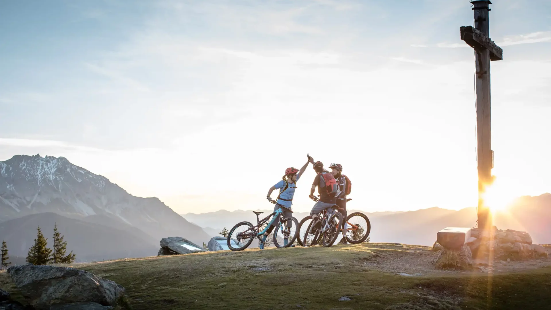 Mountainbiker feiern Gipfelsieg bei Sonnenuntergang am Bergkreuz mit Panoramablick in den Alpen