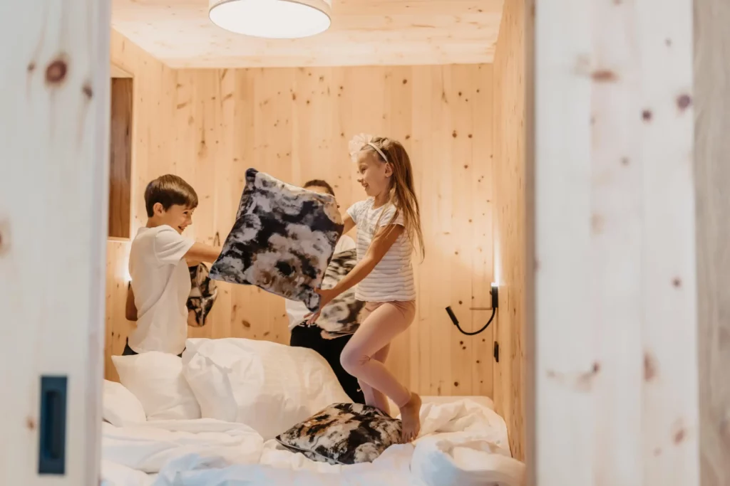 Children play happily on a bed in a cosy room with wooden panelling - cushions fly through the air in an exuberant cushion fight.