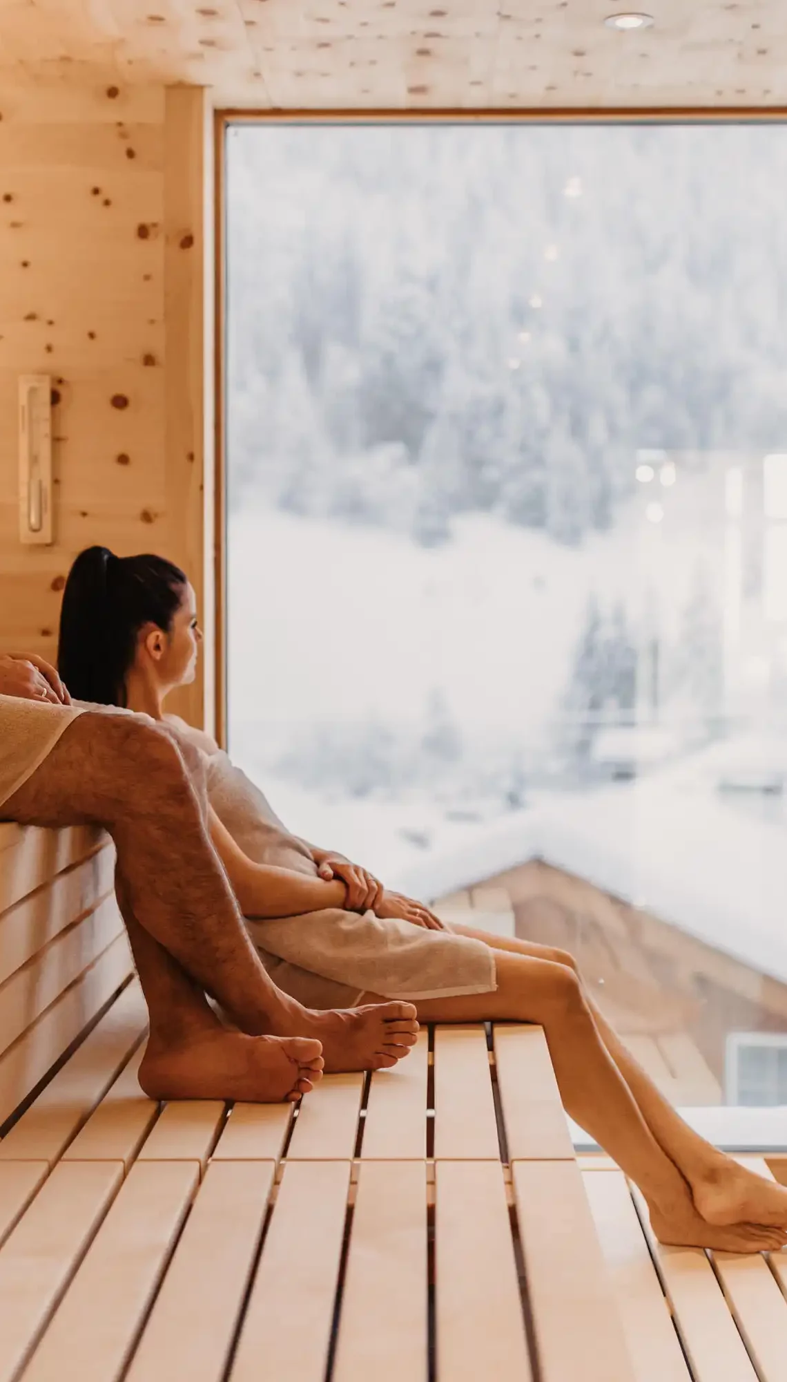 Wohlfühlmoment mit Panoramablick: Ein Paar genießt die wohltuende Wärme der Skysauna im Hofgut Wagrain mit Blick auf die verschneiten Dächer der alpinen Winterlandschaft.