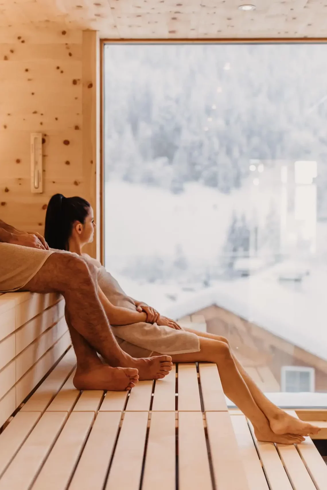 Wohlfühlmoment mit Panoramablick: Ein Paar genießt die wohltuende Wärme der Skysauna im Hofgut Wagrain mit Blick auf die verschneiten Dächer der alpinen Winterlandschaft.