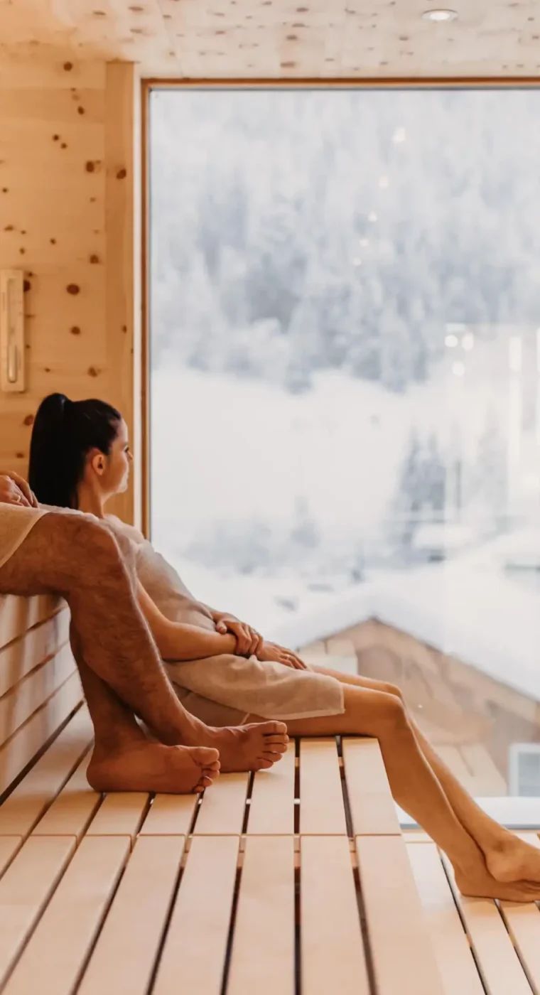 Wohlfühlmoment mit Panoramablick: Ein Paar genießt die wohltuende Wärme der Skysauna im Hofgut Wagrain mit Blick auf die verschneiten Dächer der alpinen Winterlandschaft.