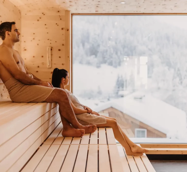 Wohlfühlmoment mit Panoramablick: Ein Paar genießt die wohltuende Wärme der Skysauna im Hofgut Wagrain mit Blick auf die verschneiten Dächer der alpinen Winterlandschaft.