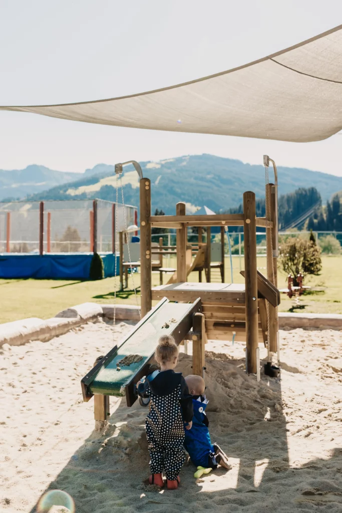 Hofgut_Spielplatz Zwei Kleinkinder spielen im Sandbereich eines Outdoor-Spielplatzes mit Holzgerüst und Wasserrinne – schattiger Bereich mit Blick auf grüne Wiesen und Berge im Hintergrund.