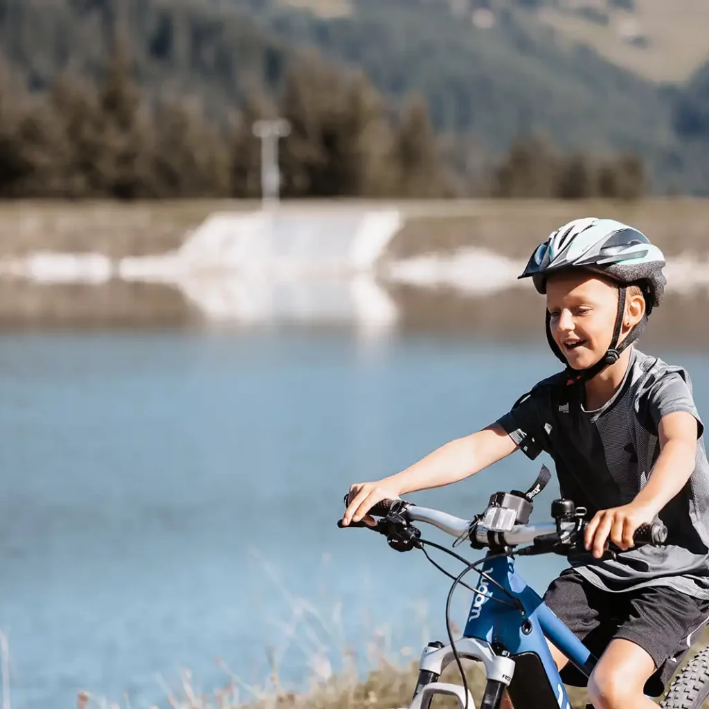 A boy in a grey shirt and bicycle helmet rides along the shore of a lake on a blue mountain bike, laughing.