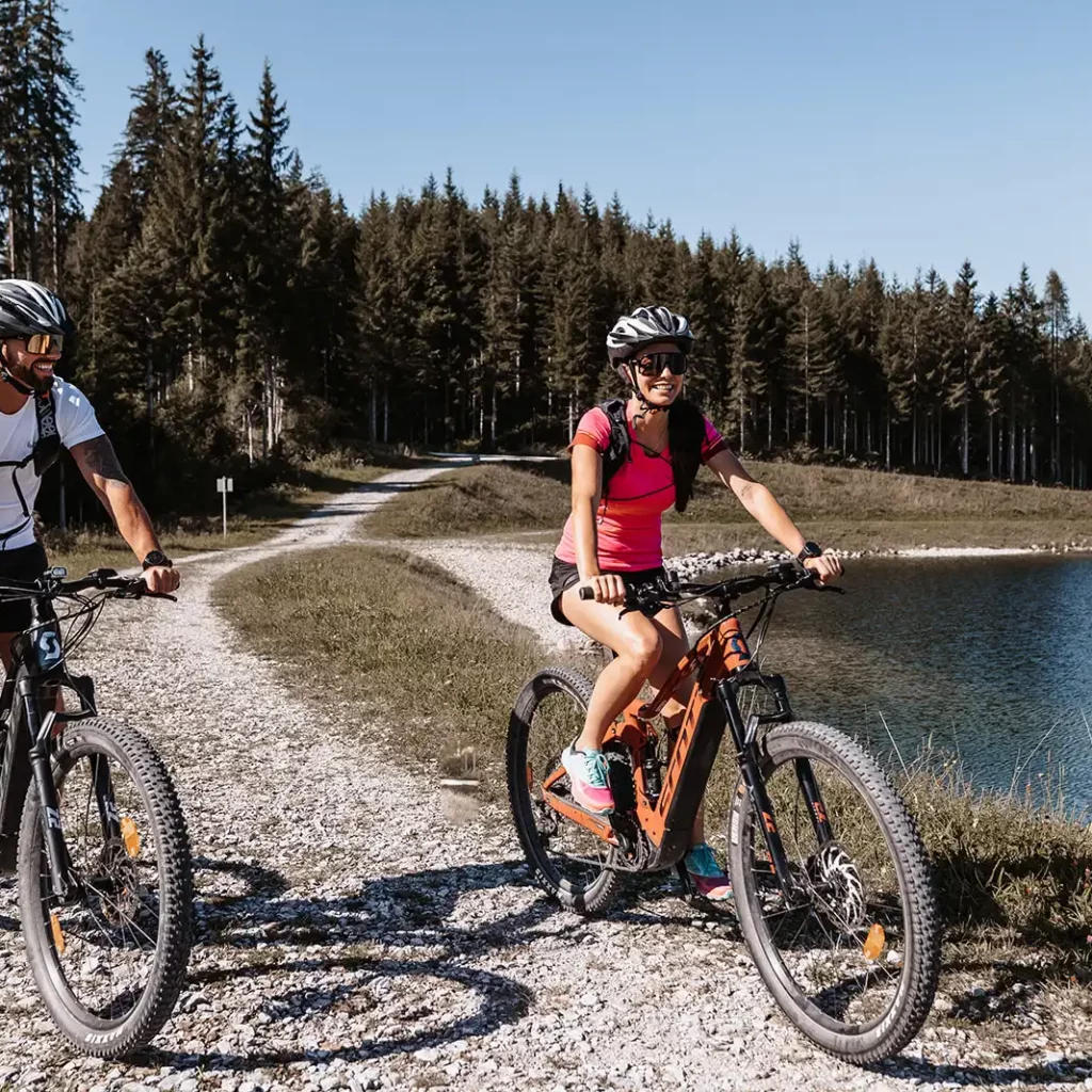A man and a woman wearing cycle helmets ride along a winding gravel track on mountain bikes, with a dense forest and lake in the background.