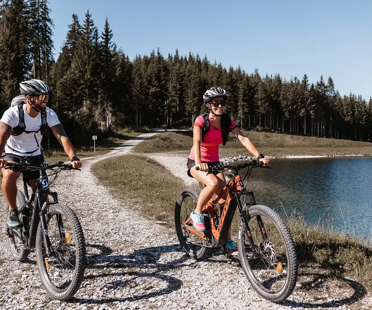 Ein Mann und eine Frau mit Fahrradhelmen fahren auf Mountainbikes einen kurvigen Schotterweg entlang, mit dichtem Wald und See im Hintergrund.