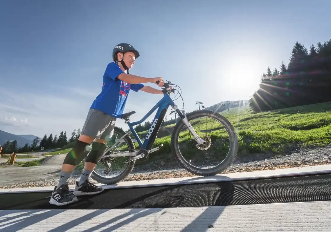 Junge mit Mountainbike auf Förderband im Bikepark bei Sonnenschein im Salzburger Land