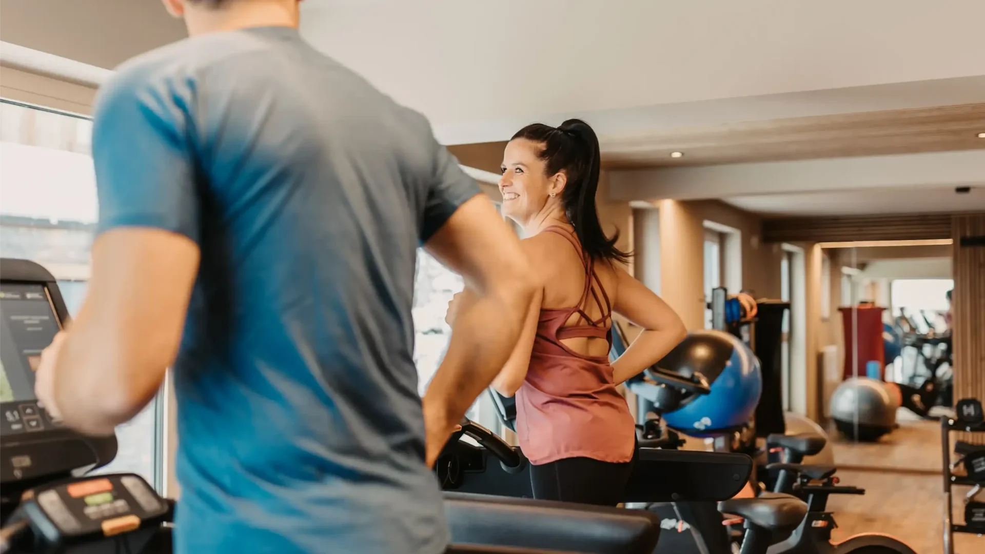Frau und Mann trainieren an modernen Fitnessgeräten im lichtdurchfluteten Gym mit Holzboden und gemütlicher Atmosphäre.