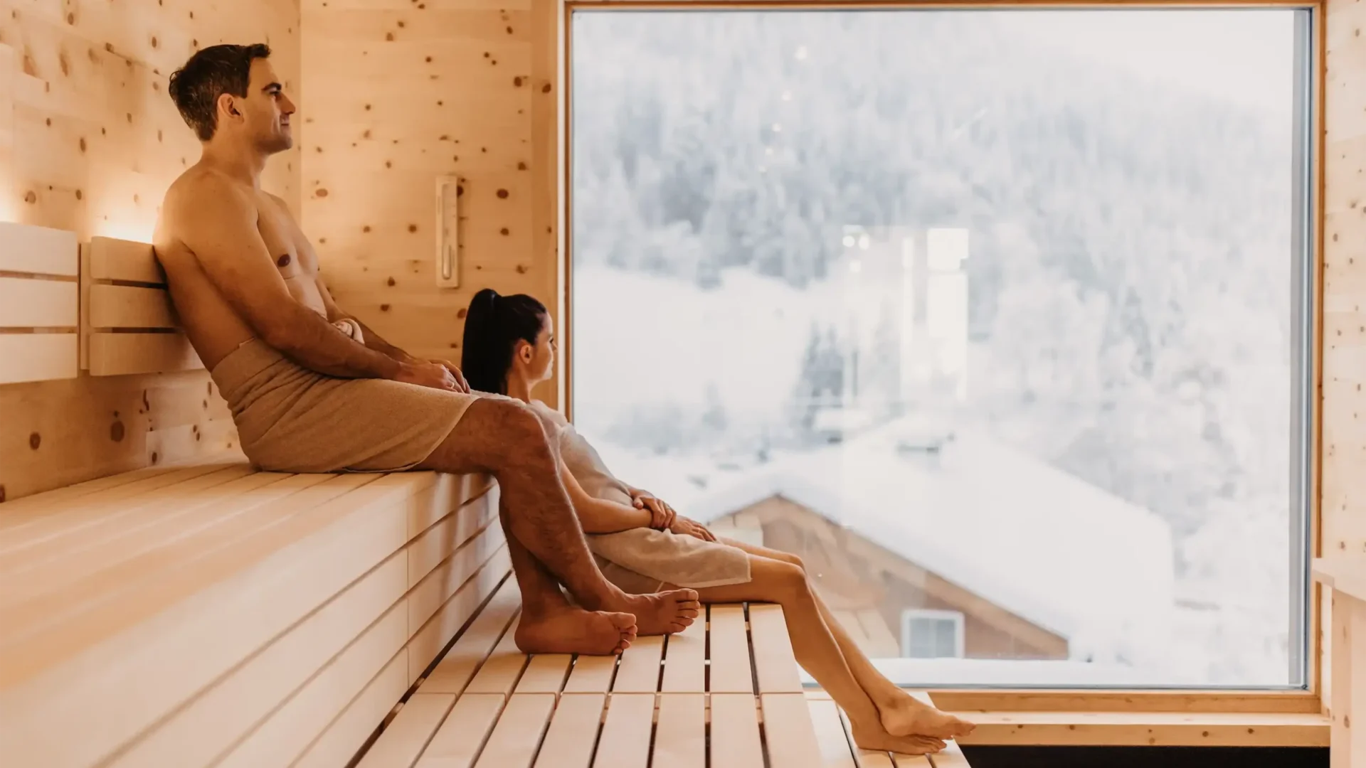Wohlfühlmoment mit Panoramablick: Ein Paar genießt die wohltuende Wärme der Skysauna im Hofgut Wagrain mit Blick auf die verschneiten Dächer der alpinen Winterlandschaft.