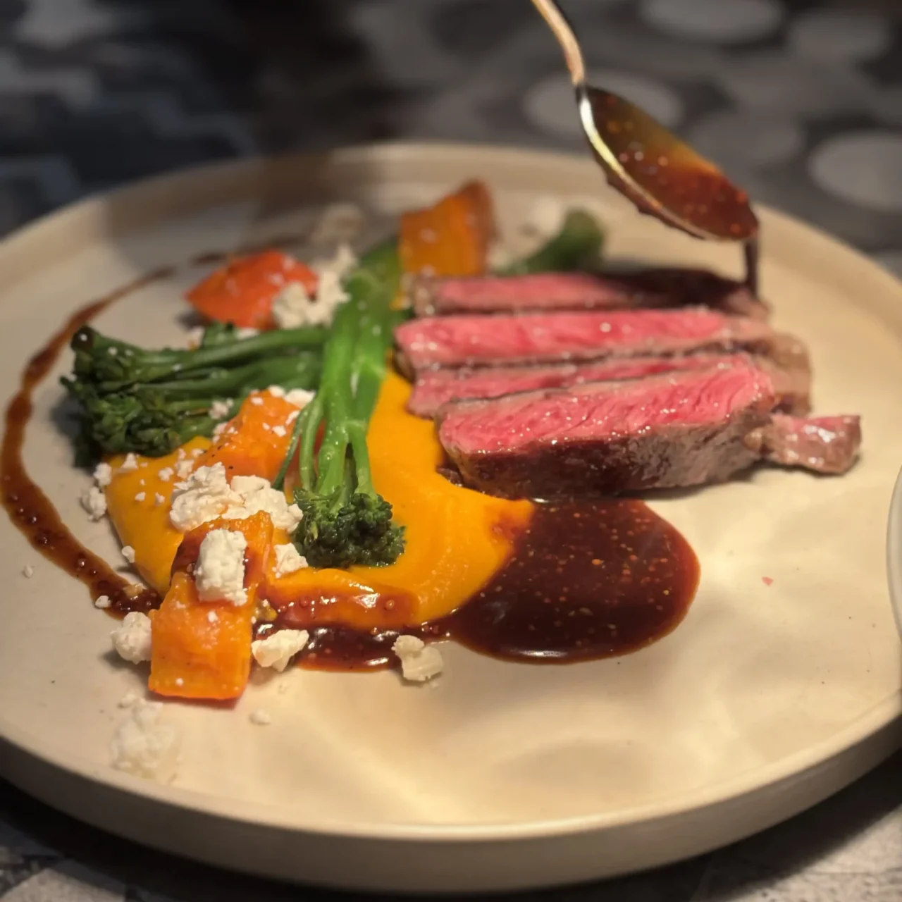 Zart rosa gebratenes Rindersteak mit Brokkolini, Kürbispüree, ofengeröstetem Kürbis und dunkler Jus, garniert mit zerbröckeltem Käse – kunstvoll angerichtet auf einem hellen Teller.