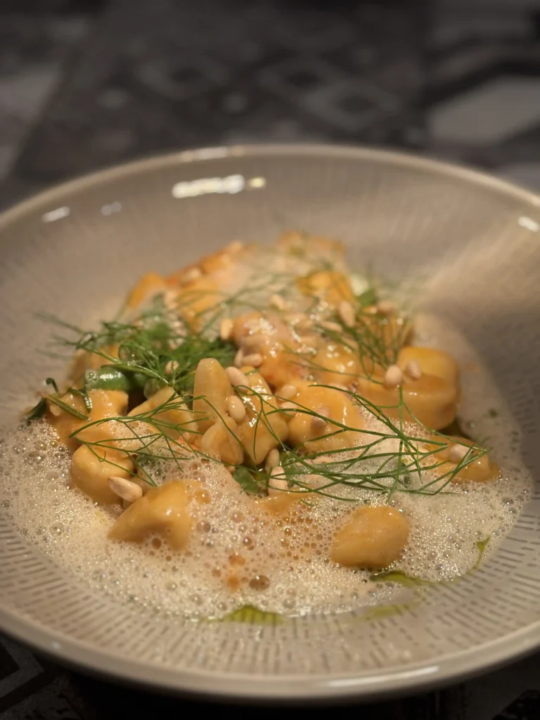 Hausgemachte Gnocchi in cremiger Sauce, verfeinert mit Schaum, frischem Dillgrün und gerösteten Pinienkernen, kunstvoll angerichtet in einem tiefen Teller.