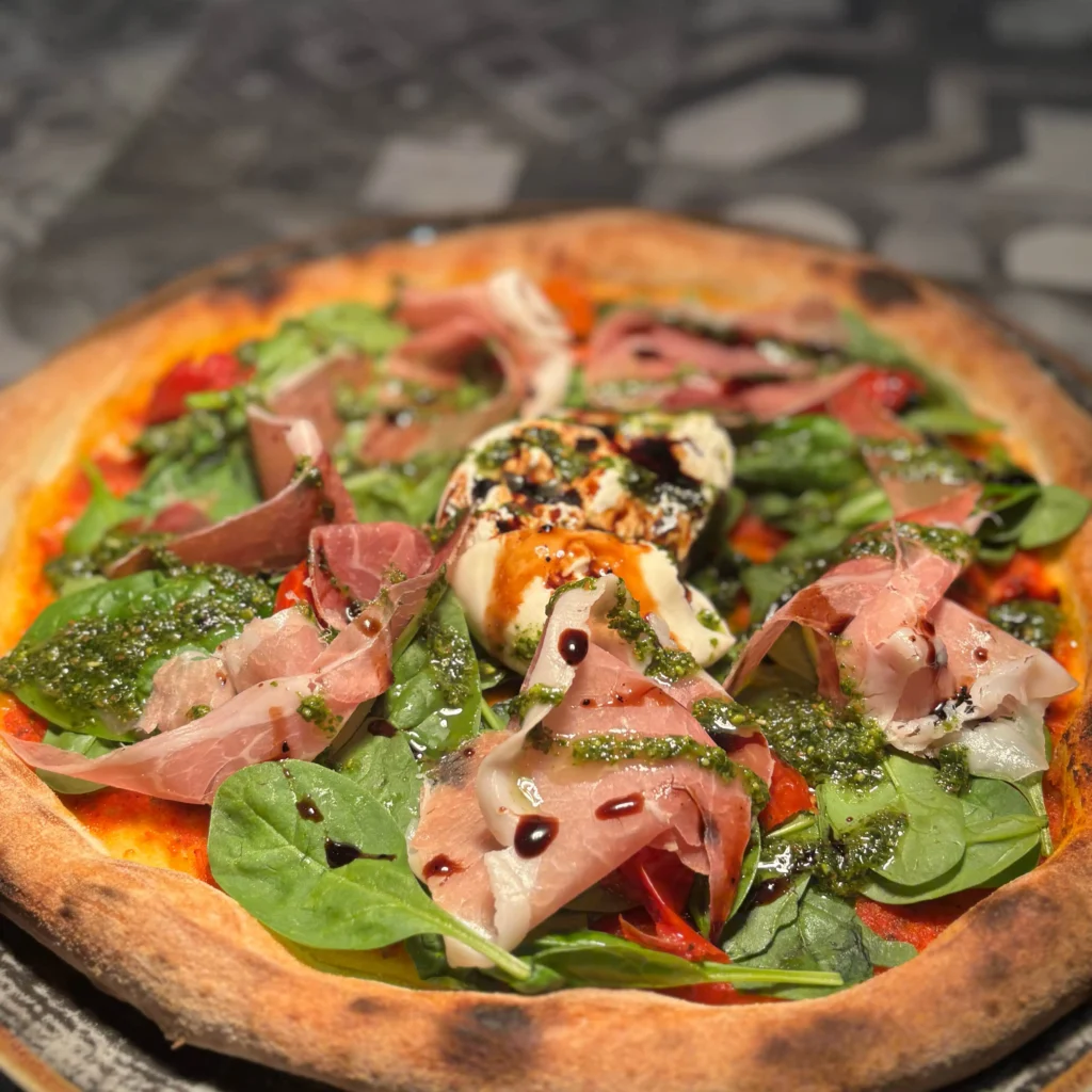 Frisch gebackene Pizza mit knusprigem Rand, belegt mit Prosciutto, frischem Spinat, Burrata, grünem Pesto und einem Hauch Balsamico-Glasur.