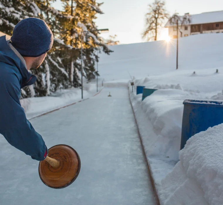 Tradition trifft Wintervergnügen – Eisstockschießen inmitten verschneiter Berglandschaft bei Sonnenuntergang.