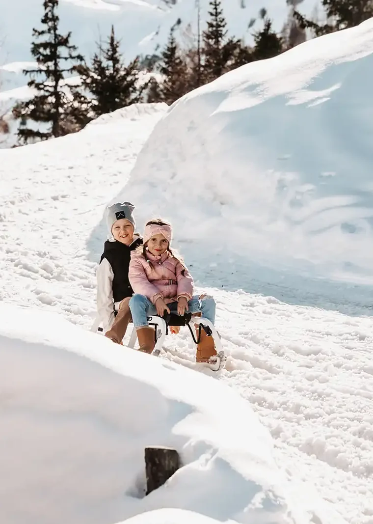 Zwei Kinder auf einem Schlitten, lachend, in Winterkleidung auf verschneitem Hang.