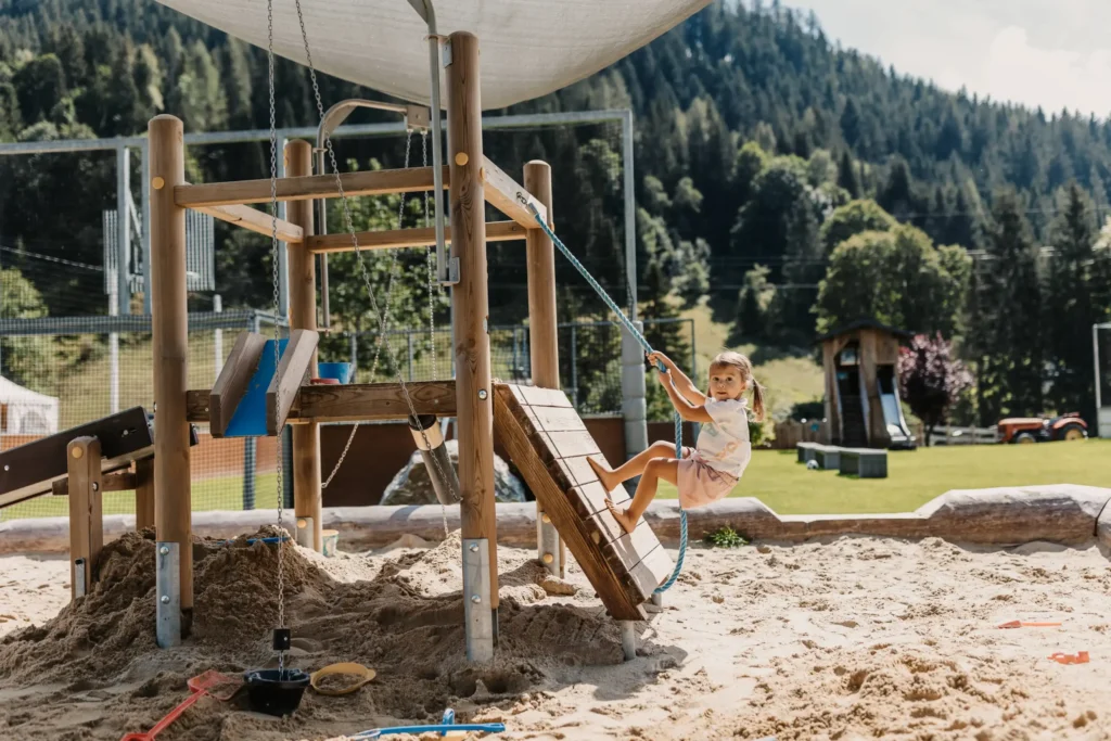 Kleines Kind spielt auf einem Holz-Klettergerüst mit Seil in einer Sandkiste am Outdoor-Spielplatz des Hofgut Wagrain, vor alpiner Bergkulisse.