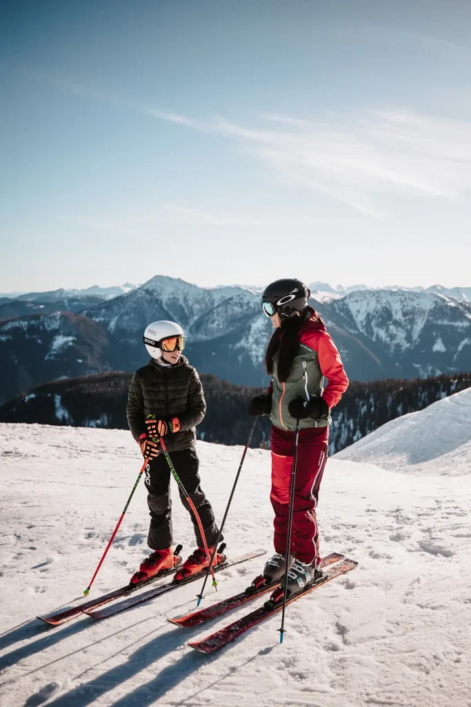 Mutter und Kind in Skiausrüstung lachen gemeinsam auf der Piste vor winterlicher Bergkulisse.